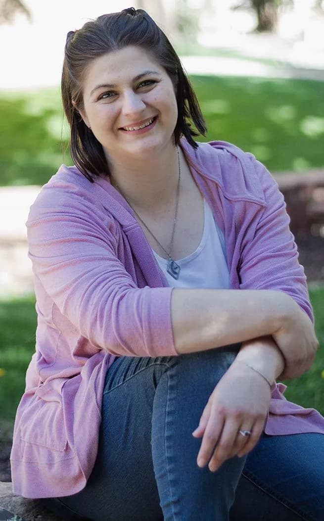 Mandy sitting in a park in her white shirt, pink cardigan, and blue jeans, smiliing